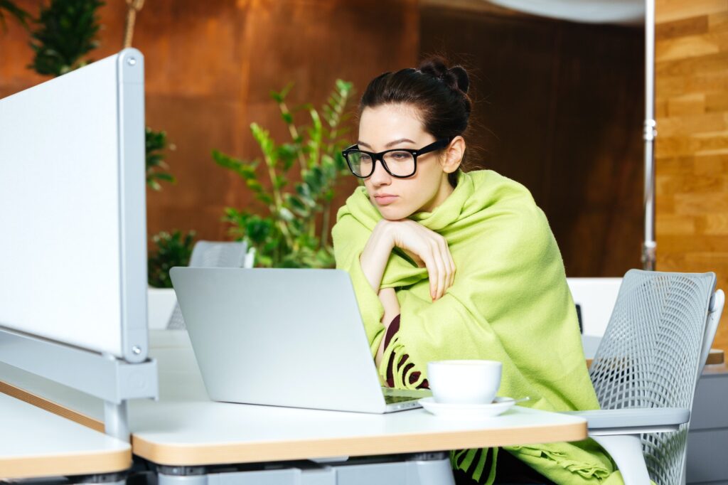 Beautiful thoughtful woman using laptop and feeling cold on workplace