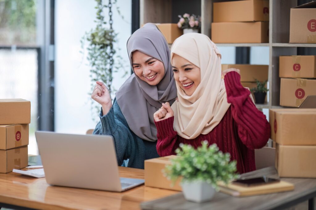 Two women muslim look at online product sales data on laptops and show joy